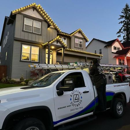 Installing Christmas lighting in Portland, OR. A 4 Seasons Softwash truck is parked in front of a grey house with lights on the roof. A worker is on the truck, preparing to install more lights.
