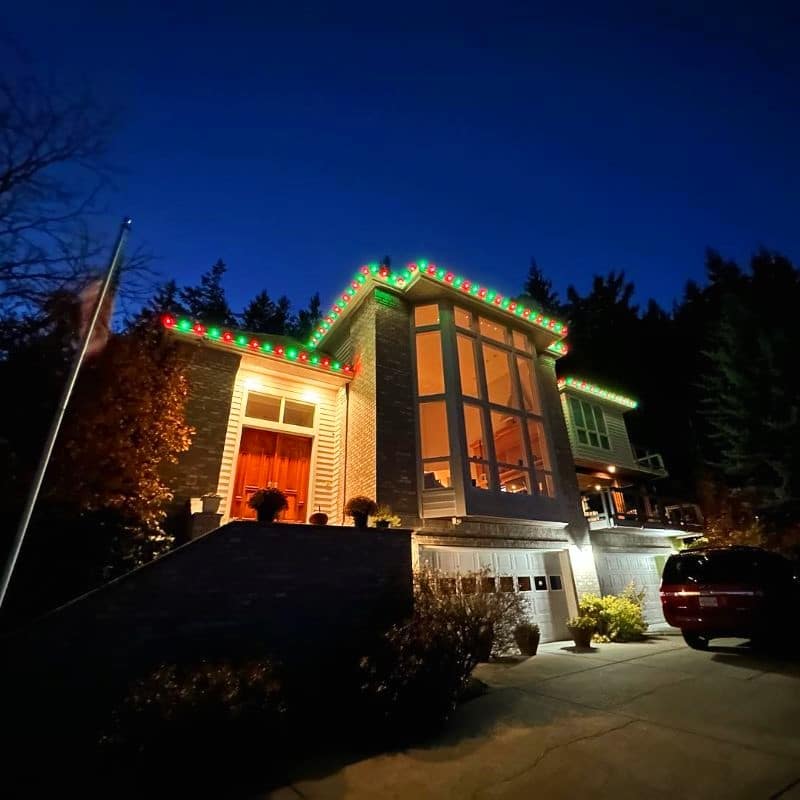 A house in Portland, OR, adorned with red and green Christmas lighting along the roofline at dusk. The large windows glow warmly, casting light onto the driveway and a parked car.