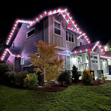 A two-story house is adorned with festive red and white Christmas lights outlining the roofline. The house is surrounded by a green lawn and various trees and bushes, all illuminated by the holiday lighting.
