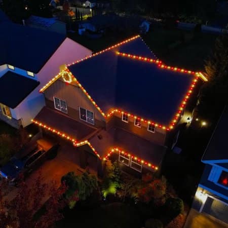 A house illuminated with warm Christmas lights outlining the roof and windows, creating a festive glow in the evening. A wreath adorns the front, adding to the holiday cheer.