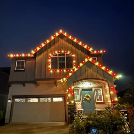A two-story house is decorated with bright red and yellow Christmas lights outlining the roof and windows. A wreath hangs on the front door, and a security sign is visible in the yard.