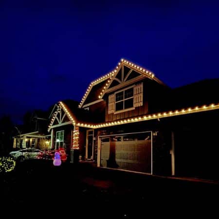 A home in Portland, OR, adorned with warm white Christmas lighting outlining its roofline and gables. A glowing snowman and festive decorations add to the holiday cheer.
