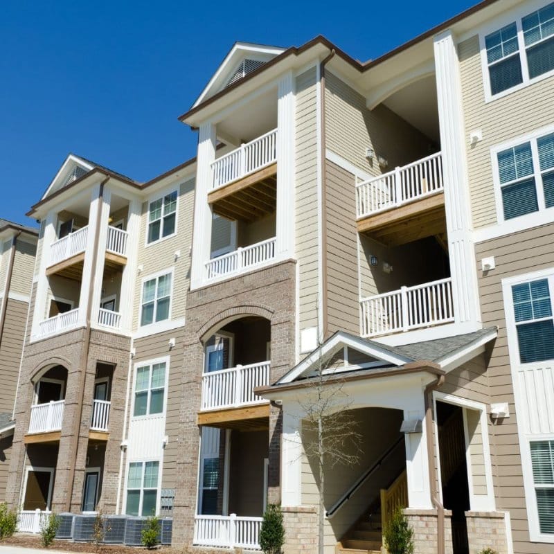 Modern, multi-story apartment building with tan siding, brick accents, and white balconies against a clear blue sky. Ideal for commercial building washing projects.