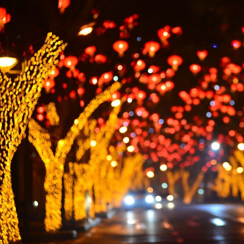 Night view of a street lined with trees illuminated with yellow lights, and red lanterns hanging above, creating a festive atmosphere for Commercial Christmas Lighting.