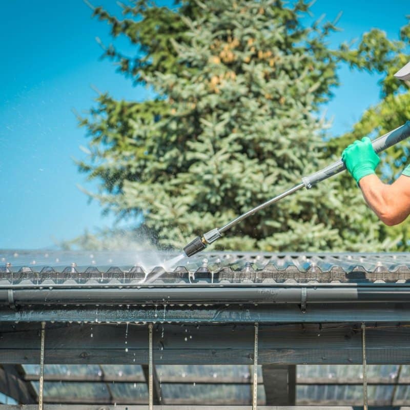 Gutter cleaning in Portland OR: A worker wearing green gloves pressure washes gutters with a backdrop of a lush green tree and a bright blue sky.