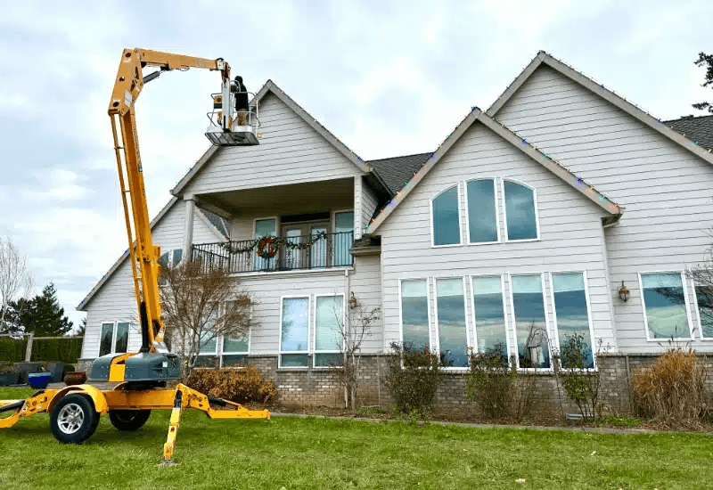 A yellow lift elevates a worker installing Christmas lights on a large, two-story home. The house, possibly prepped for pressure washing, features a balcony with a wreath and many large windows reflecting the sky.