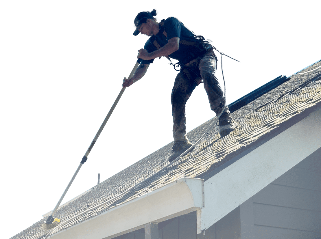 A worker pressure washing a roof. The man in safety gear uses a long-handled brush to clean moss and debris from the shingles, showcasing home maintenance.