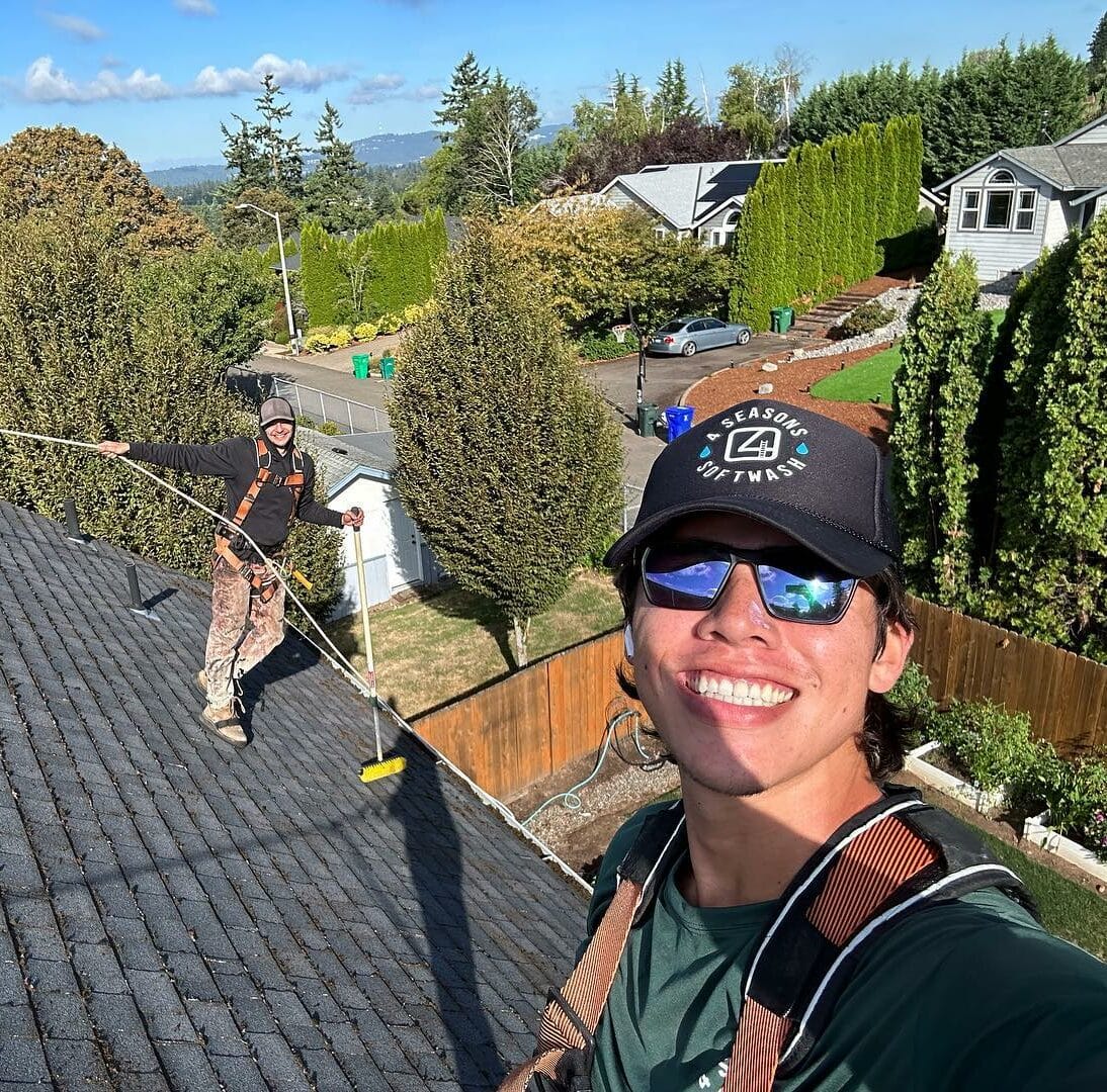 Two workers on a roof are doing pressure washing. One wears a "4 Seasons Softwash" hat. Houses and trees are in the background.