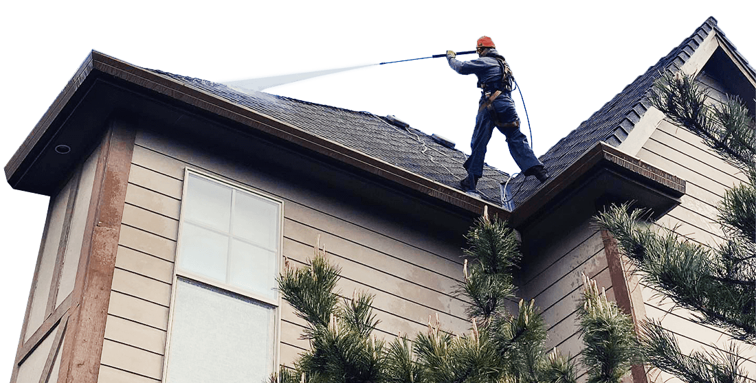 Professional roof cleaning in Portland, OR. A worker in safety gear pressure washes a shingled roof, removing dirt and debris. The house has tan siding and brown trim.