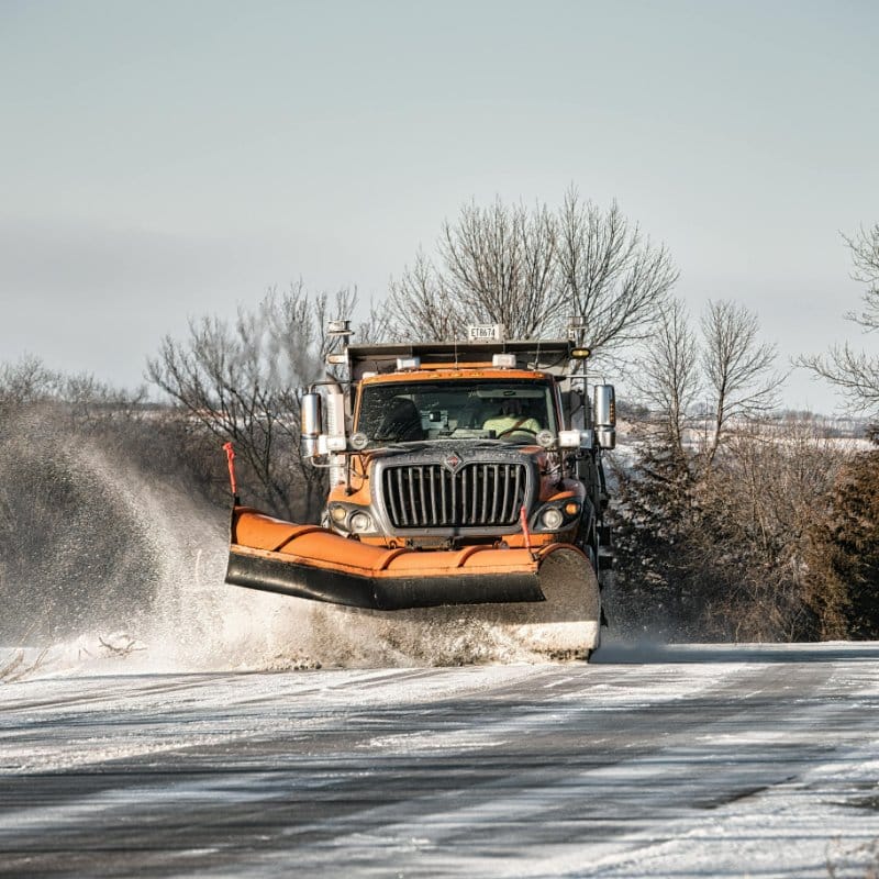 Orange snow plow truck clearing a road, spraying snow and water to the sides. Winter scene with bare trees; snow removal in progress.