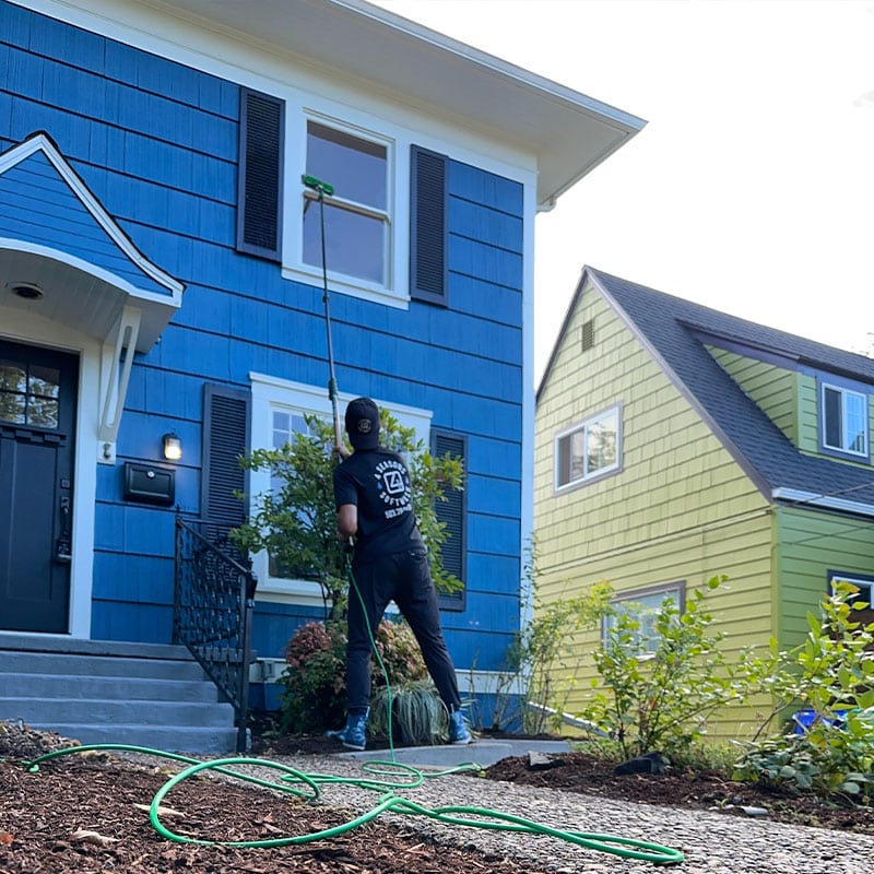 A window washing professional in Portland OR uses an extension pole to clean the second-story window of a blue house with black shutters. A green hose lies on the ground. A yellow house is visible in the background.