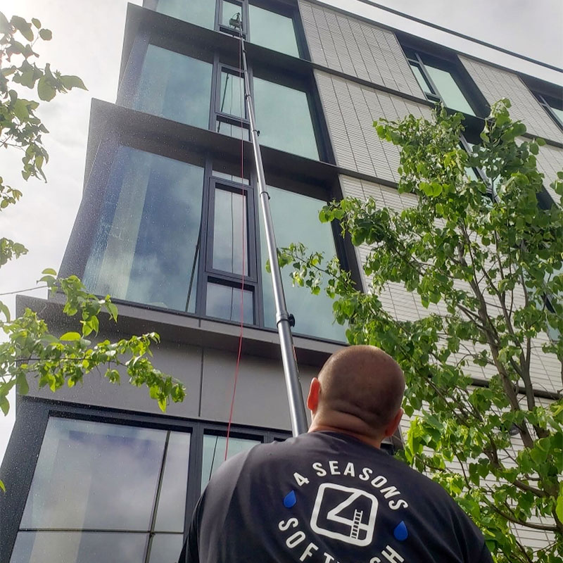 A window cleaner uses a long pole to clean the tall windows of a modern commercial building in Portland, OR. The worker's shirt reads "4 Seasons Soft Wash". Commercial window cleaning in action.