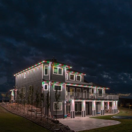 A modern two-story house illuminated with Permanent Holiday Lighting in Portland OR. Red, green, and white lights outline the roof and porch against a dramatic dark sky.
