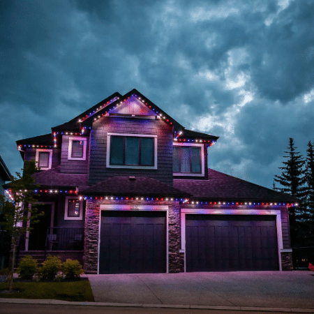 Home with permanent holiday lighting in Portland OR, showcasing red, white, and blue lights outlining the roofline and garage doors against a dramatic, cloudy evening sky.