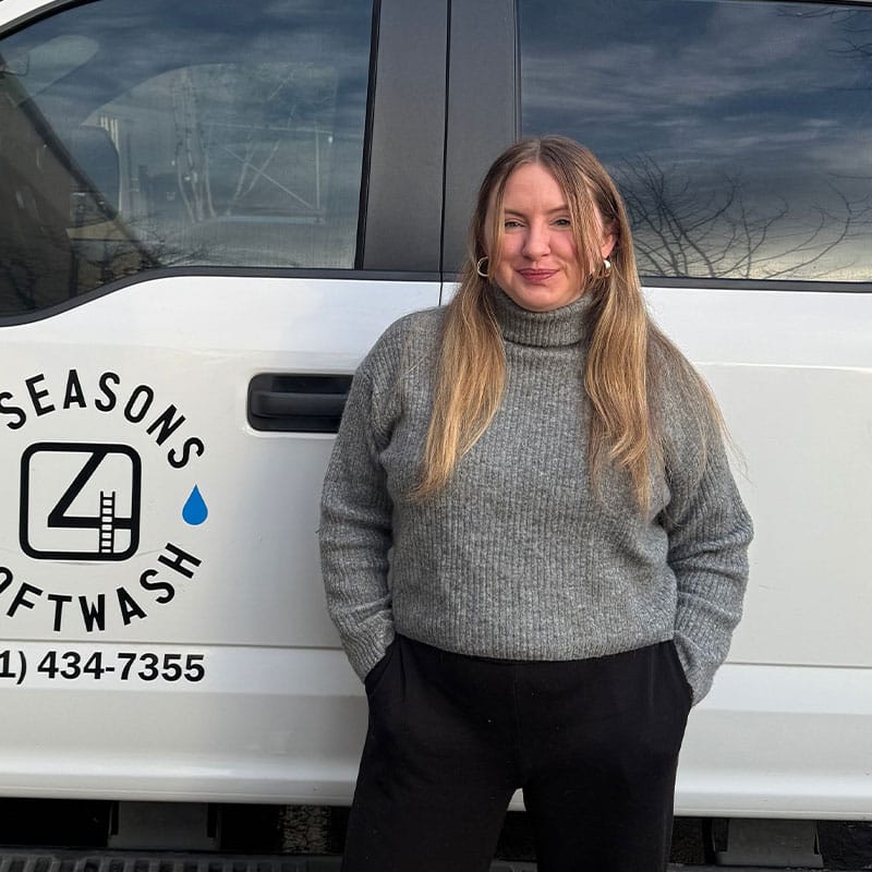 Woman stands in front of a "4 Seasons Softwash" truck. She has long blonde hair, wears a gray turtleneck and black pants, and smiles confidently. The logo and company name are prominently displayed on the truck.